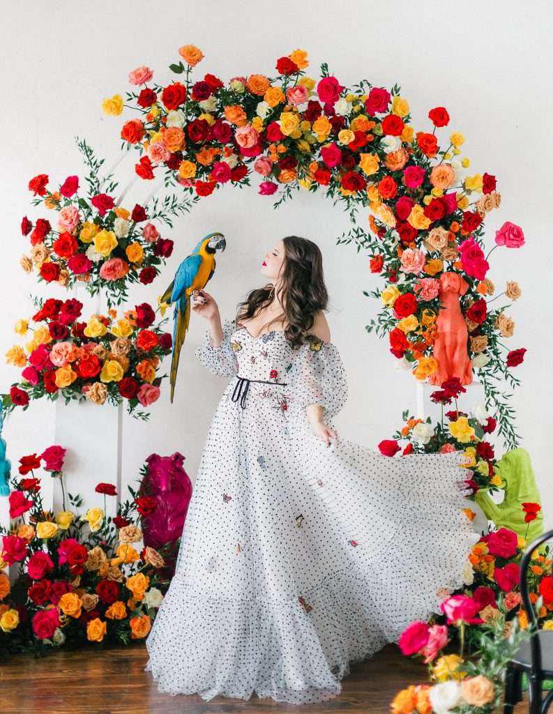 Bride in polka dot gown with a macaw under a vibrant garden rose arch