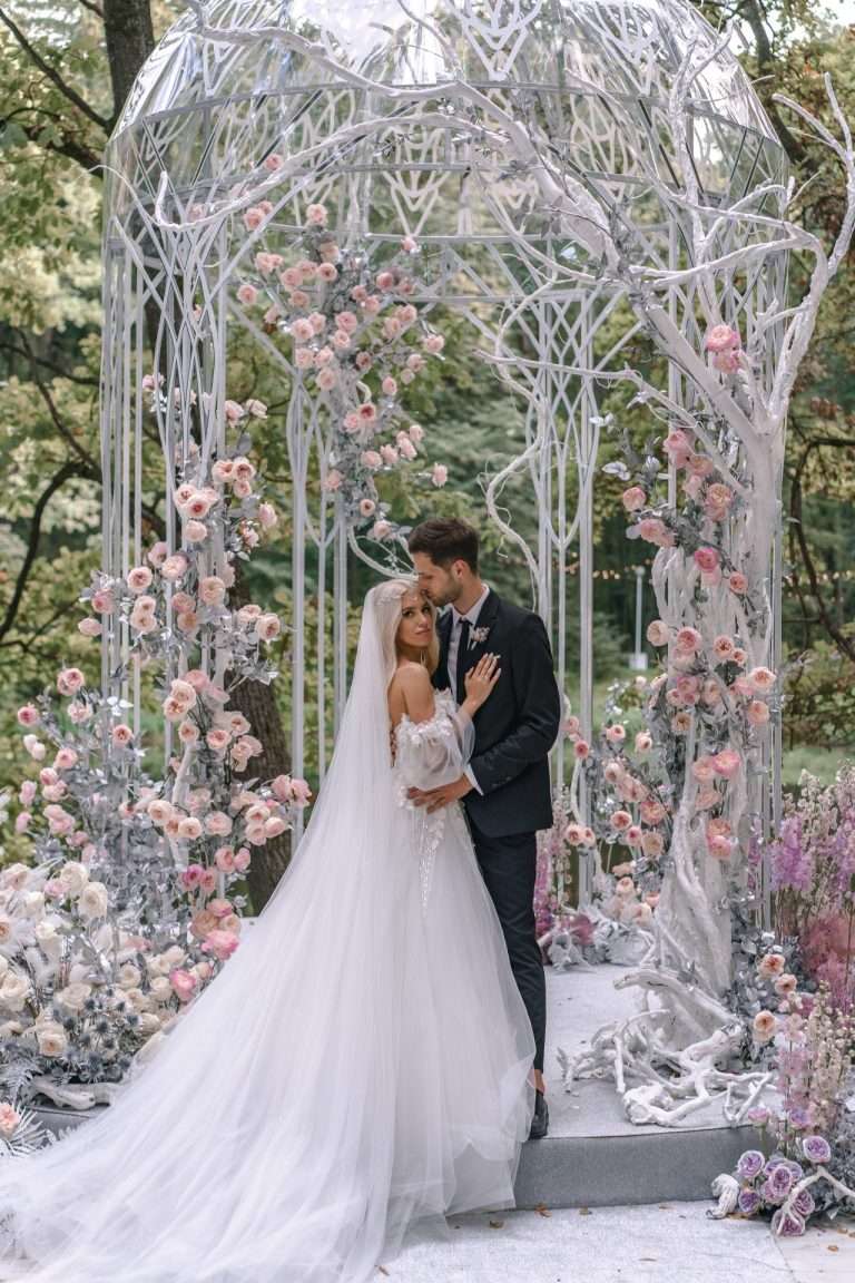 A couple embracing under a white arch covered in pink blossom.