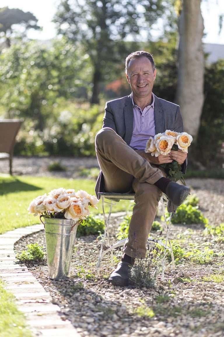 A person seated on a bench in a garden holding cream garden roses, with a bucket of blooms beside them.