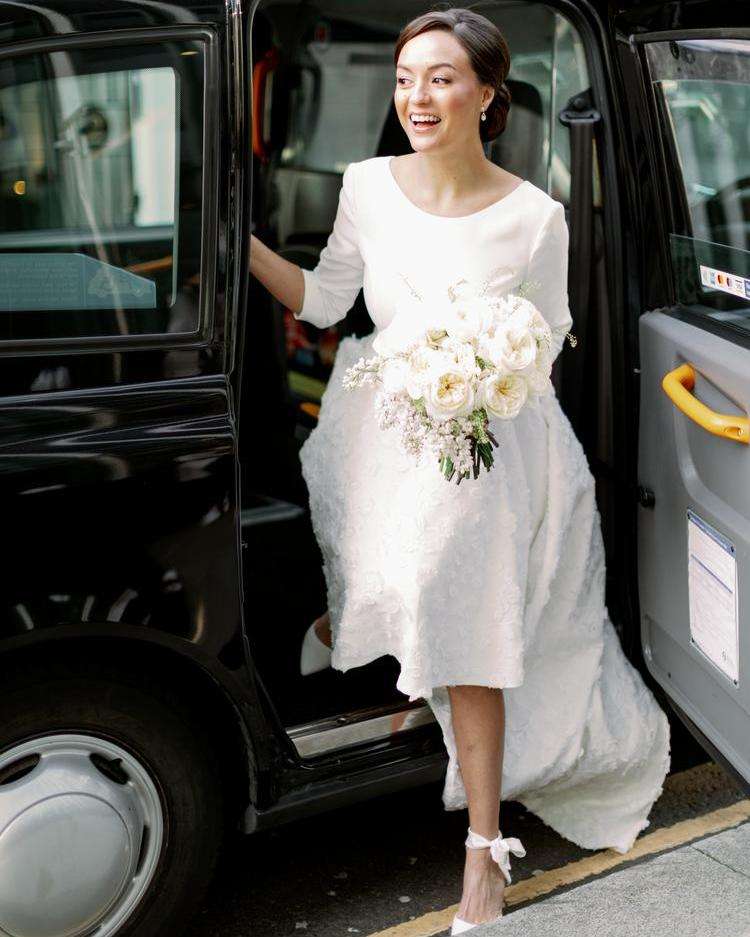 A person in a white dress stepping out of a black taxi holding a small bouquet.