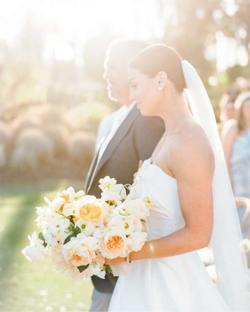 Bride and her father walking together, sunlit garden rose bouquet in hand