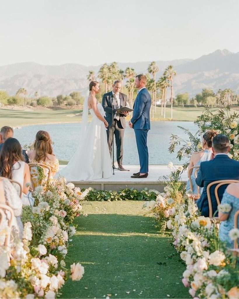 Couple exchanging vows on the lakeside stage flanked by rose-lined aisle