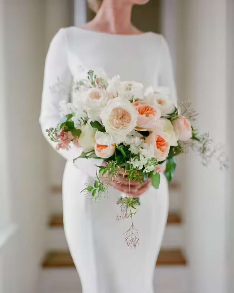 A person in a long-sleeved gown holding a bouquet of cream and peach garden roses.
