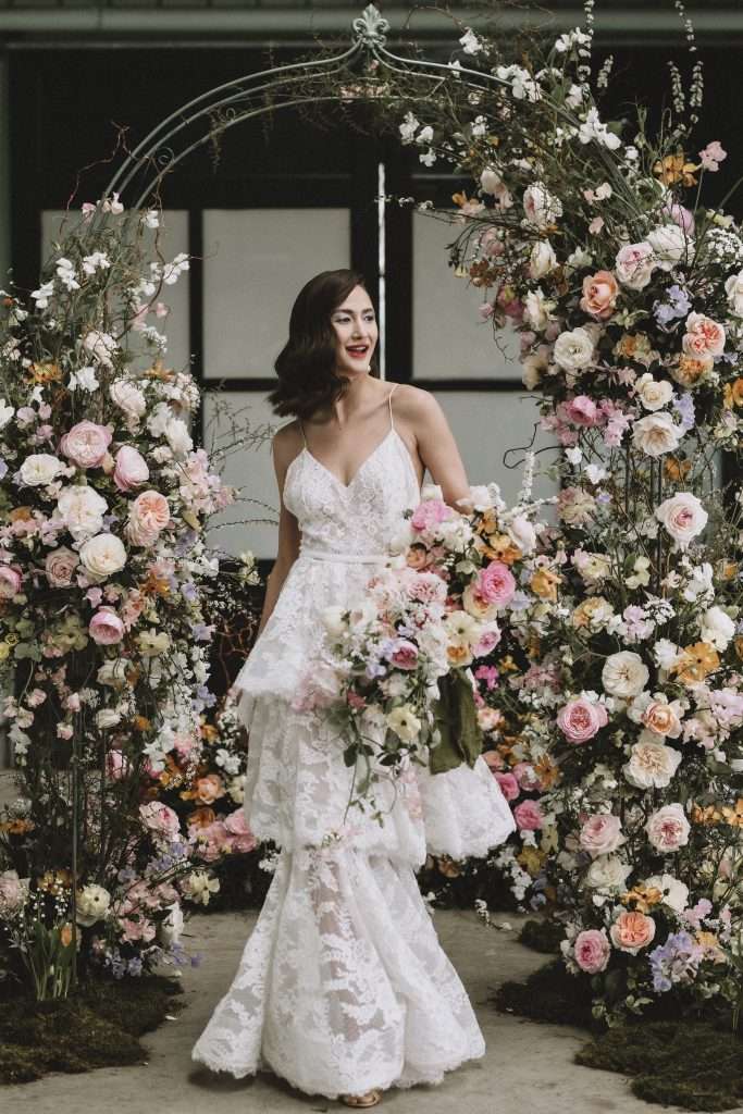 Bride under a floral arch of pink and peach garden roses, wedding work category