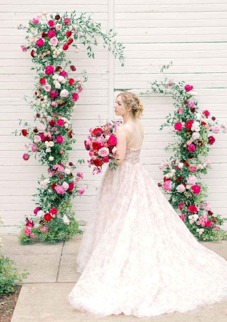 Bride holding a red and pink bouquet beside a flower-covered arch