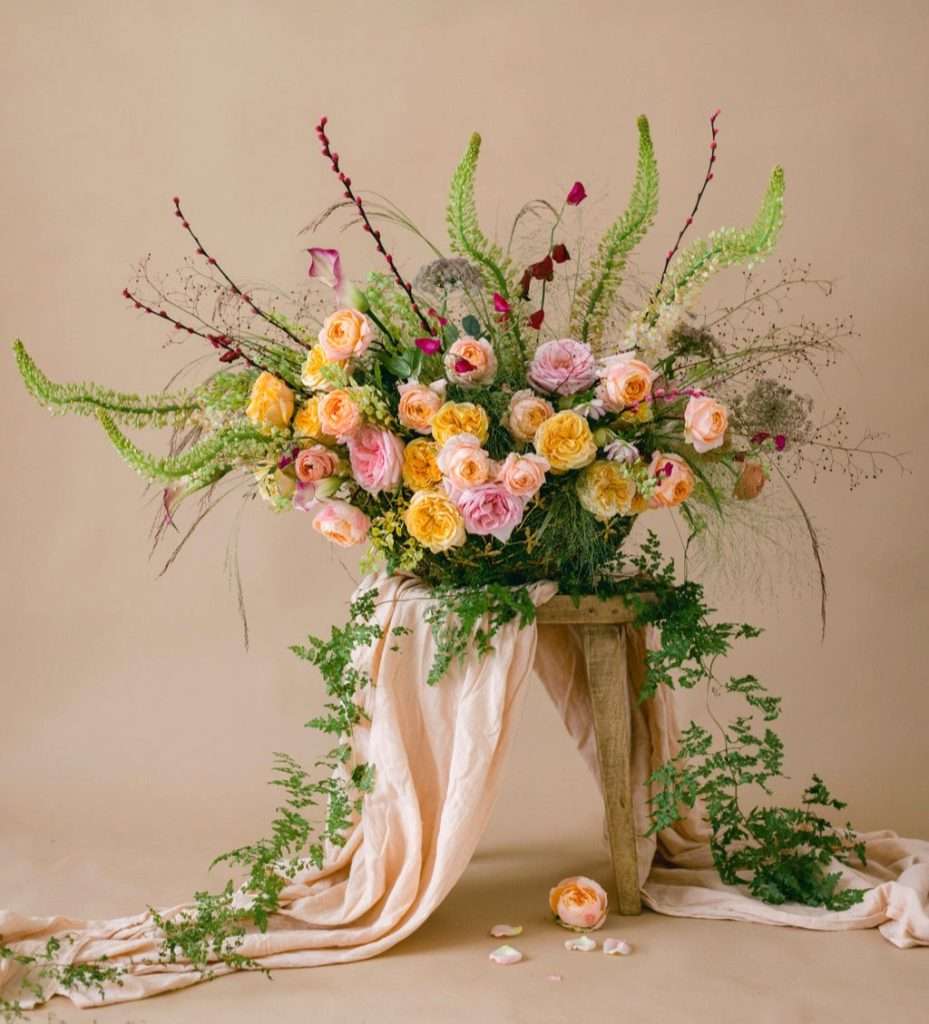 Yellow and peach garden rose arrangement with trailing greenery on a wooden stool