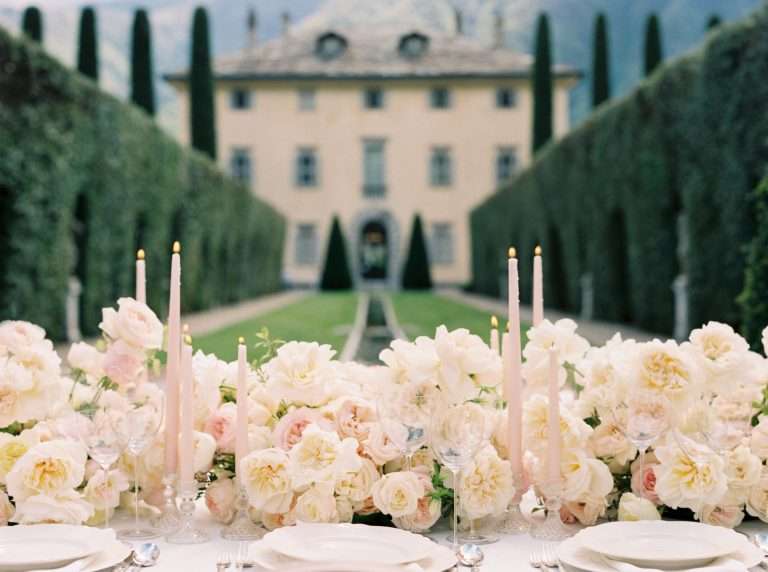 A long table set with cream garden roses and tall candles, in front of a villa and clipped hedges.