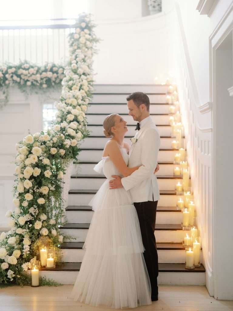 A couple embracing at the foot of a staircase framed by a white floral arch and candles.