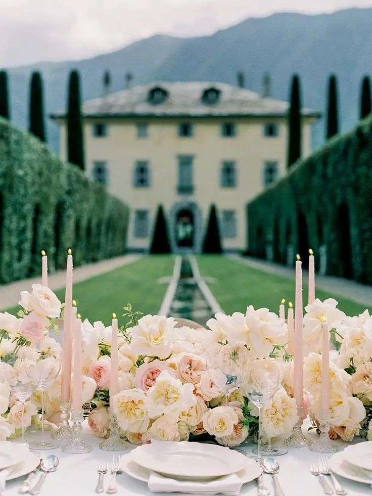 Candlelit banquet table with cream garden roses framed by hedges and a villa backdrop