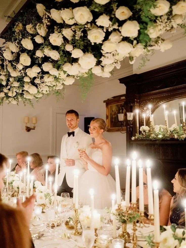 Bride and groom toasting under a suspended white garden rose floral installation at a candlelit dinner