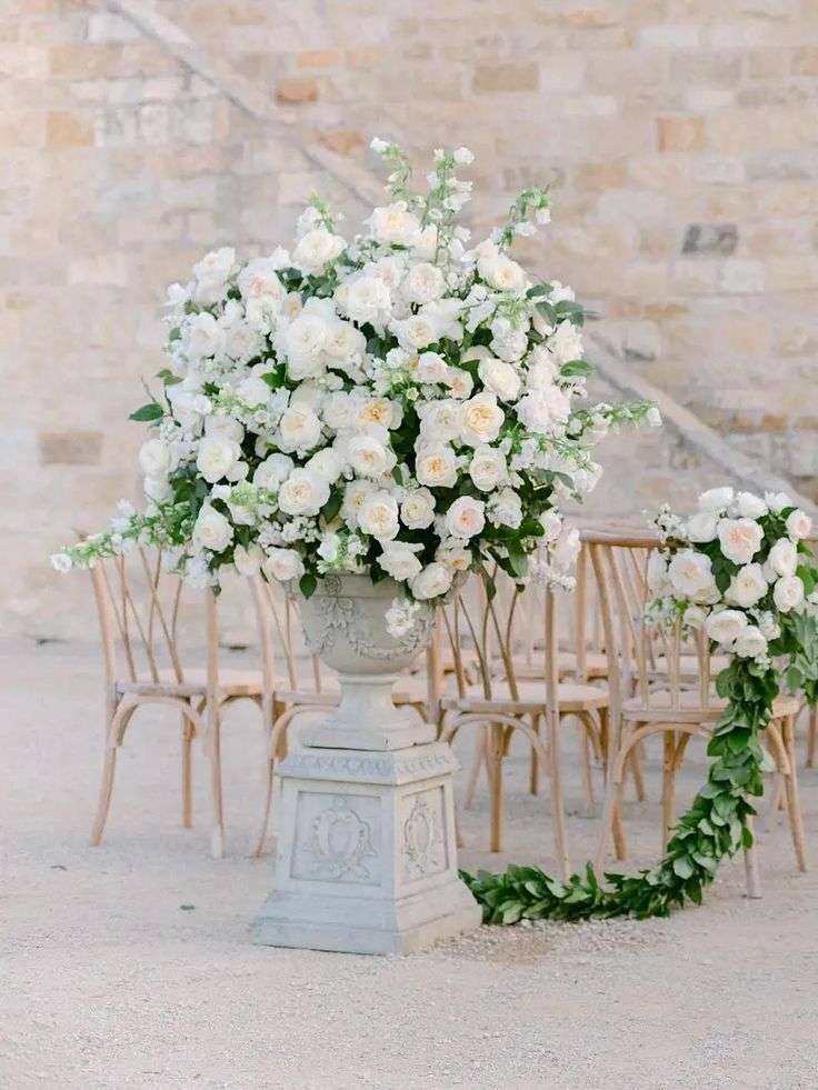 A tall urn of white garden roses and greenery beside wooden chairs at a stone venue.