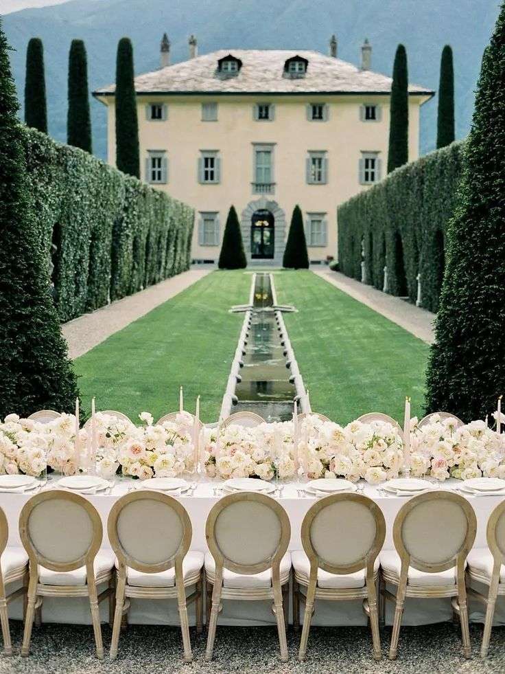 Long banquet table lined with blush garden roses facing a villa and manicured gardens