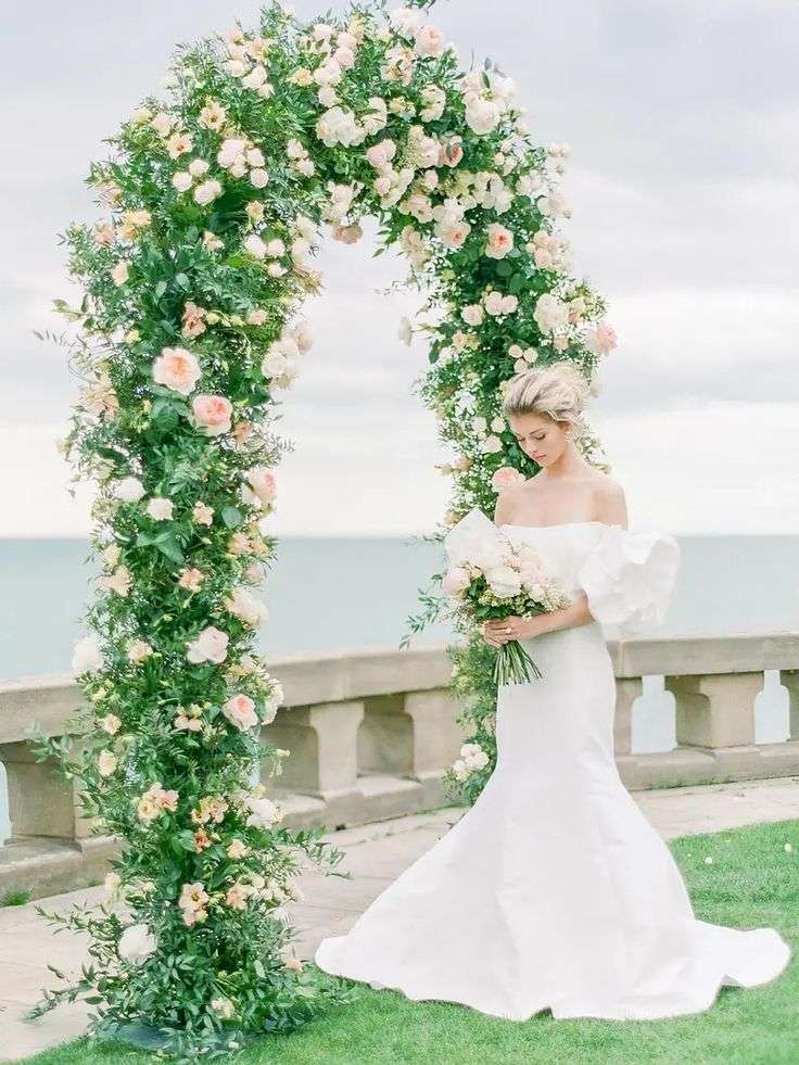 Bride in an off-shoulder gown beneath a blush and white garden rose ceremony arch by the sea