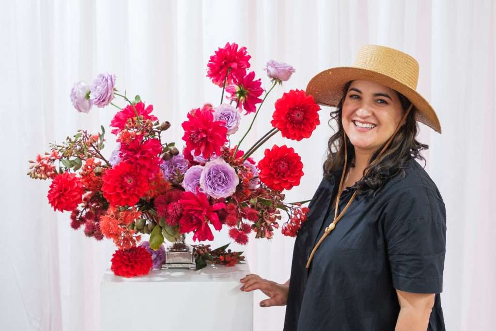 Smiling designer in a straw hat beside a red dahlia and lavender garden rose arrangement