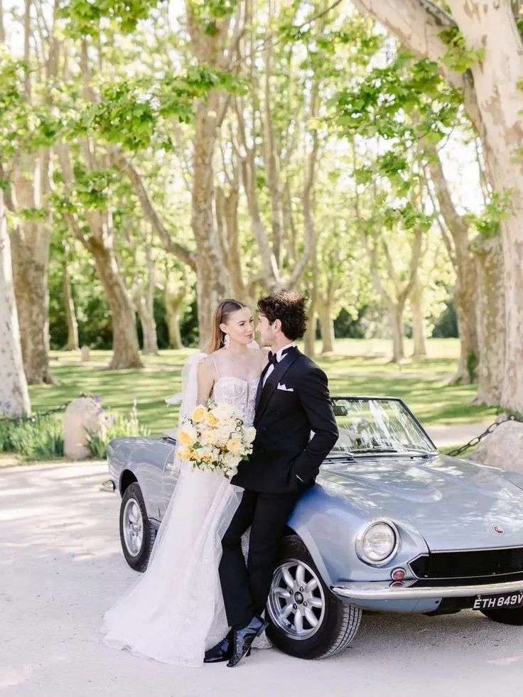 Bride and groom leaning against a vintage car under plane trees, bouquet in hand