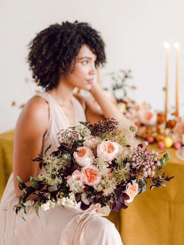 Bride seated at a gold-linen table holds a wild peach and burgundy autumn bouquet