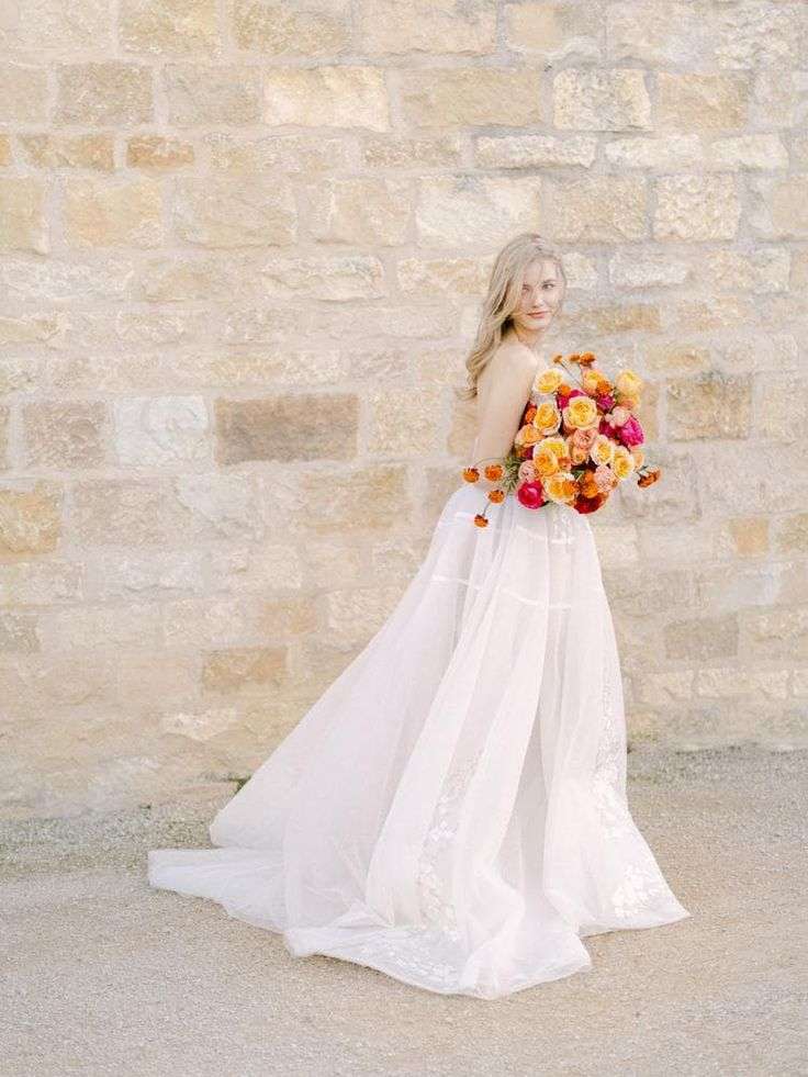 Bride in flowing gown carries a bold orange and pink bouquet against a stone wall