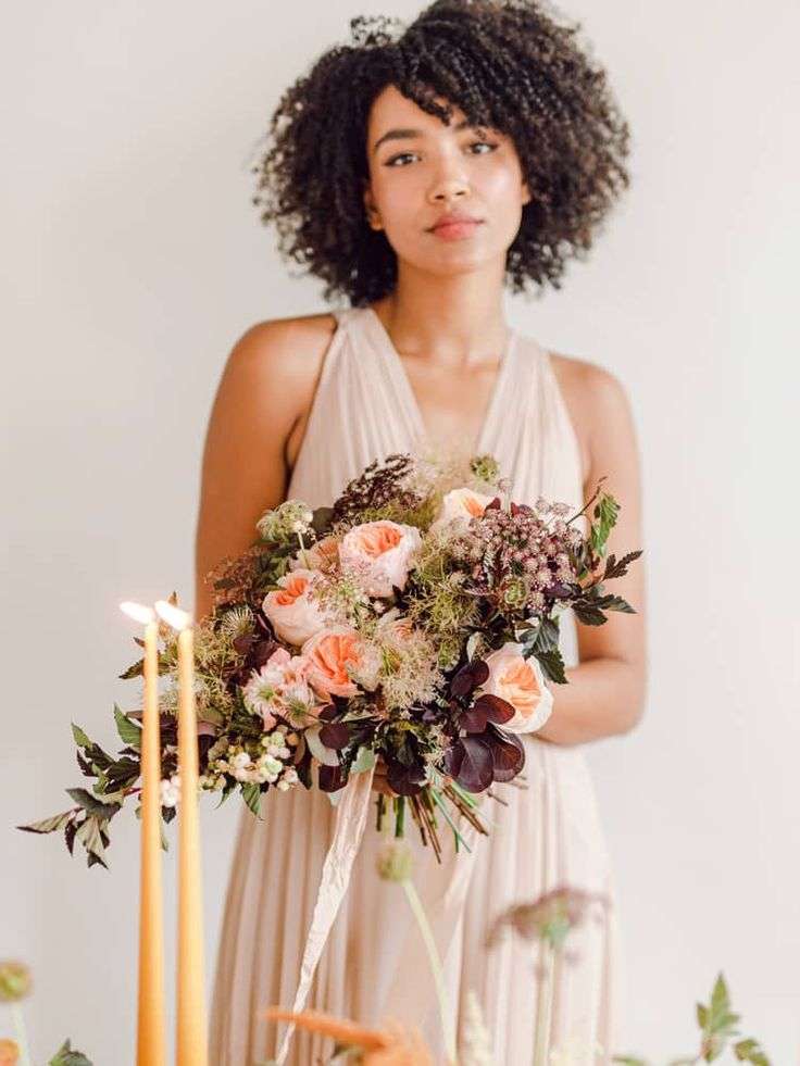 Woman in blush dress holds a moody peach and burgundy bouquet beside candlelight