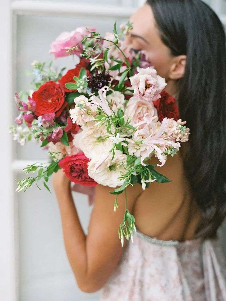 A person holding a loose bouquet of red, blush and cream garden roses up beside their face.
