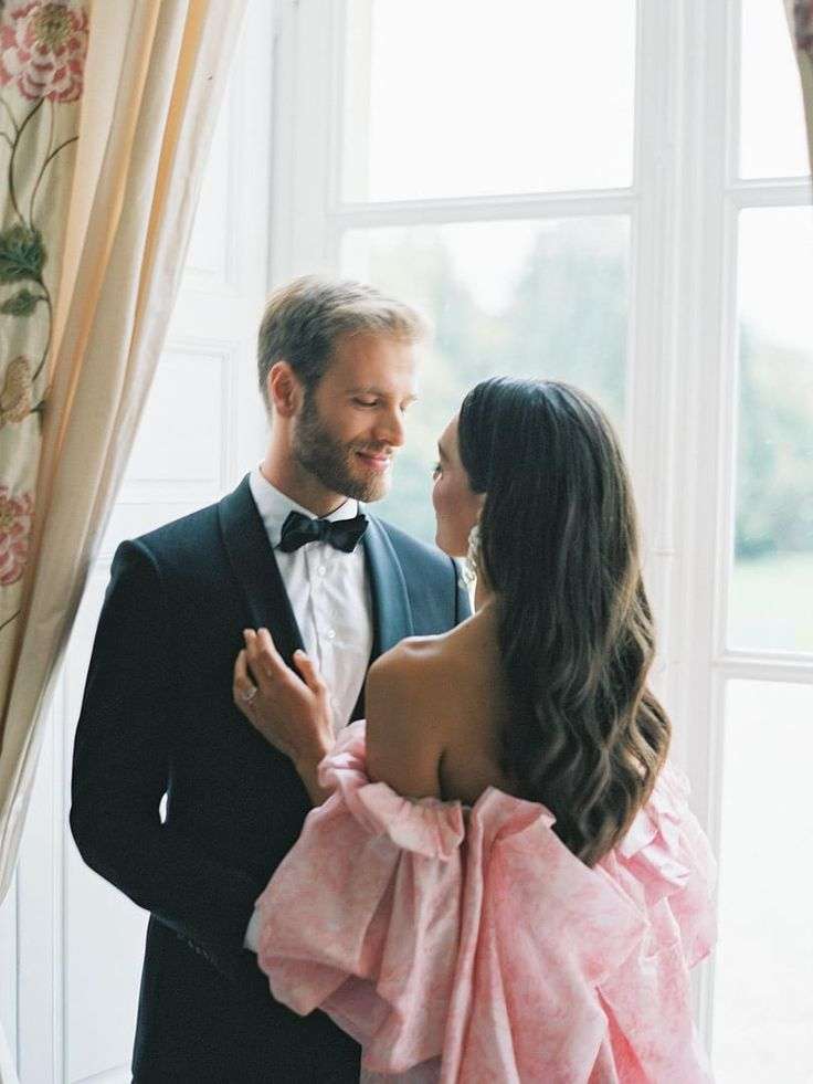 Groom in tuxedo embracing bride in ruffled pink gown by a window