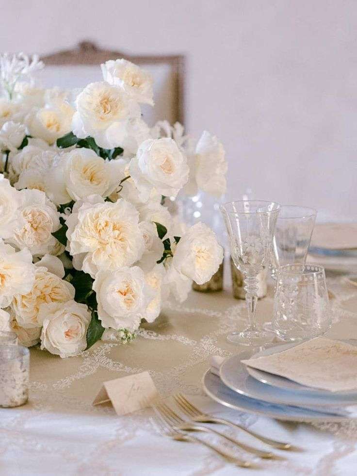 Cream-white garden rose centerpiece on a lace table with etched crystal glasses and silver place setting