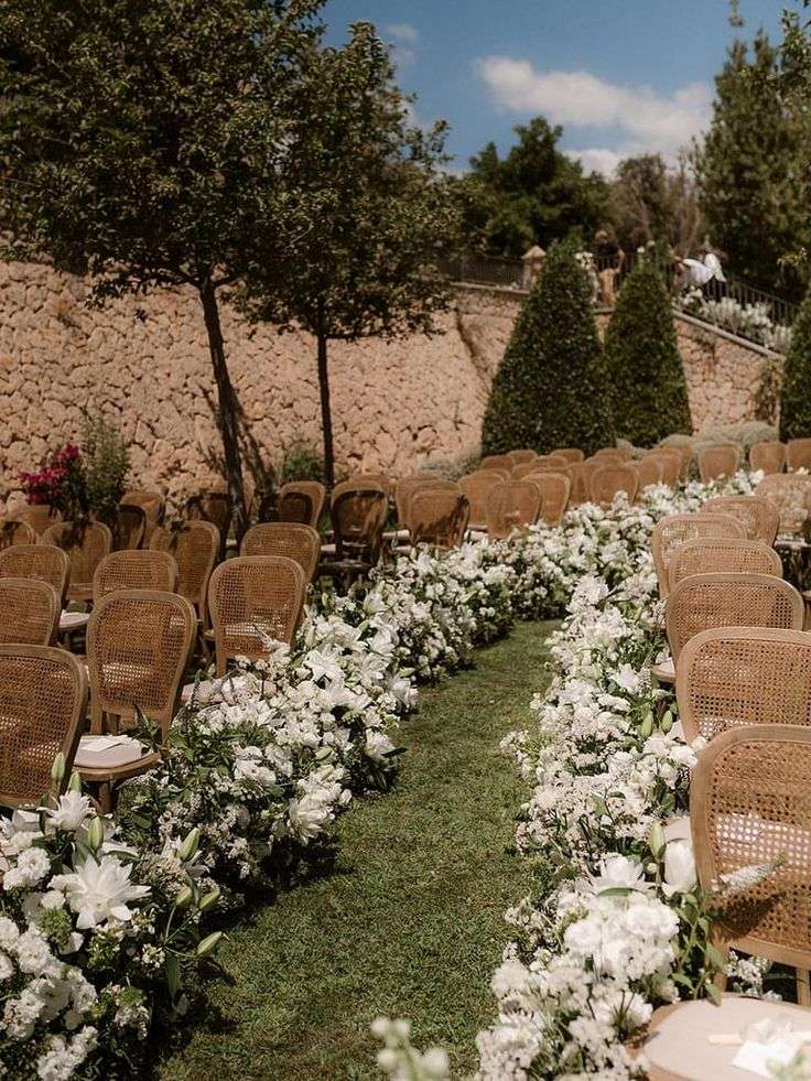 Sunlit ceremony aisle bordered with white florals along a stone garden wall