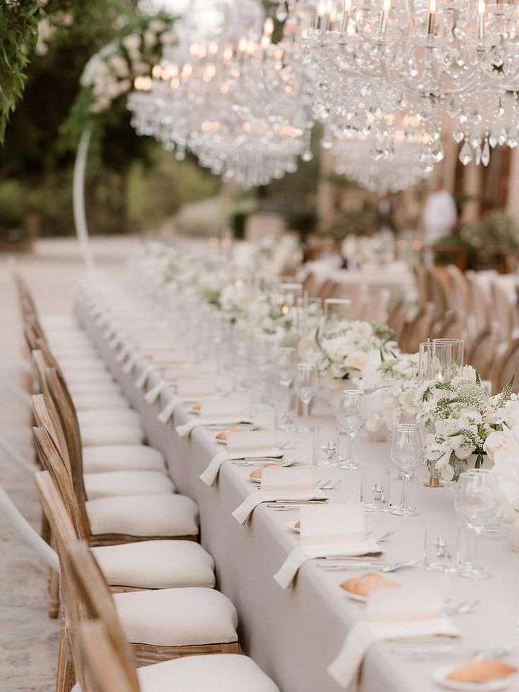 Long candlelit dinner table set under crystal chandeliers with white flowers