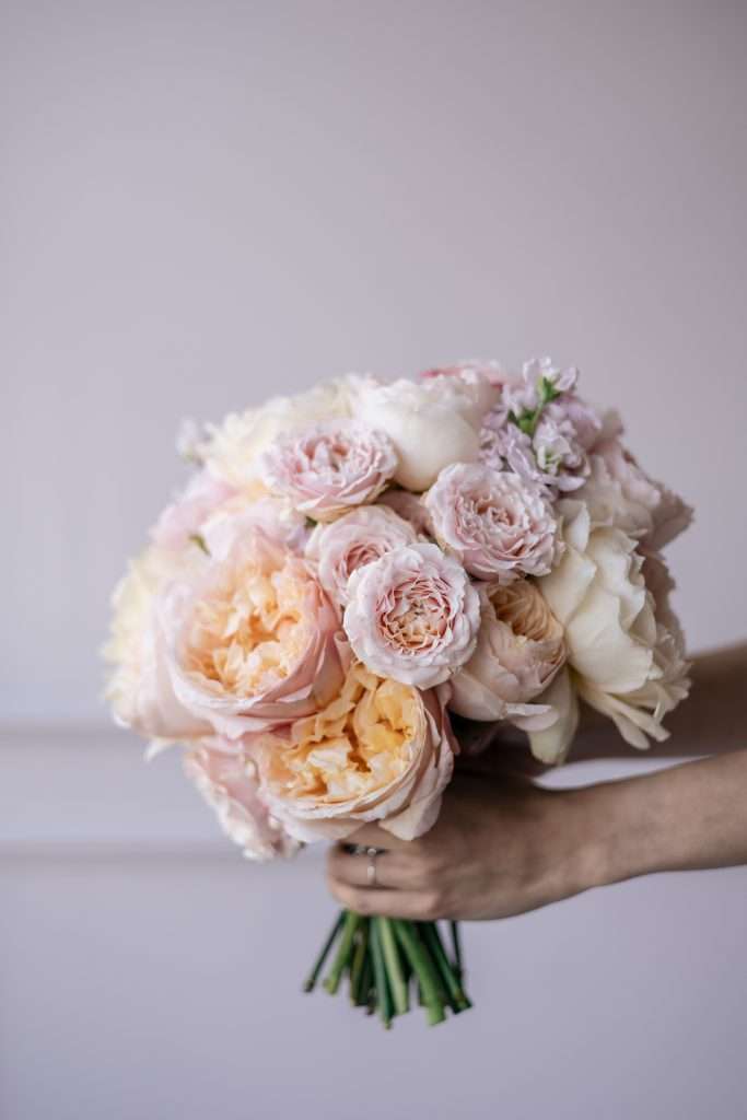 Hand holding a blush and peach garden rose bouquet against a wall