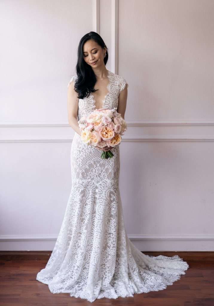 Bride in lace gown looking down at a peach-pink bouquet