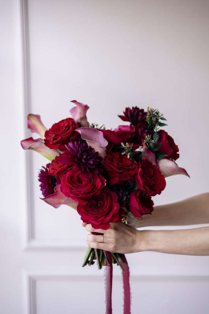 Hands presenting a bold red garden rose, dahlia and calla lily bouquet